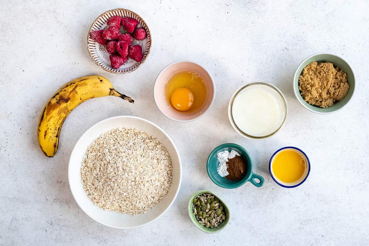 A flat lay of Healthy Baked Oats ingredients on a light surface: a banana, a bowl of oats, raspberries, an egg, milk, brown sugar, melted butter, seeds, baking powder, and cinnamon.