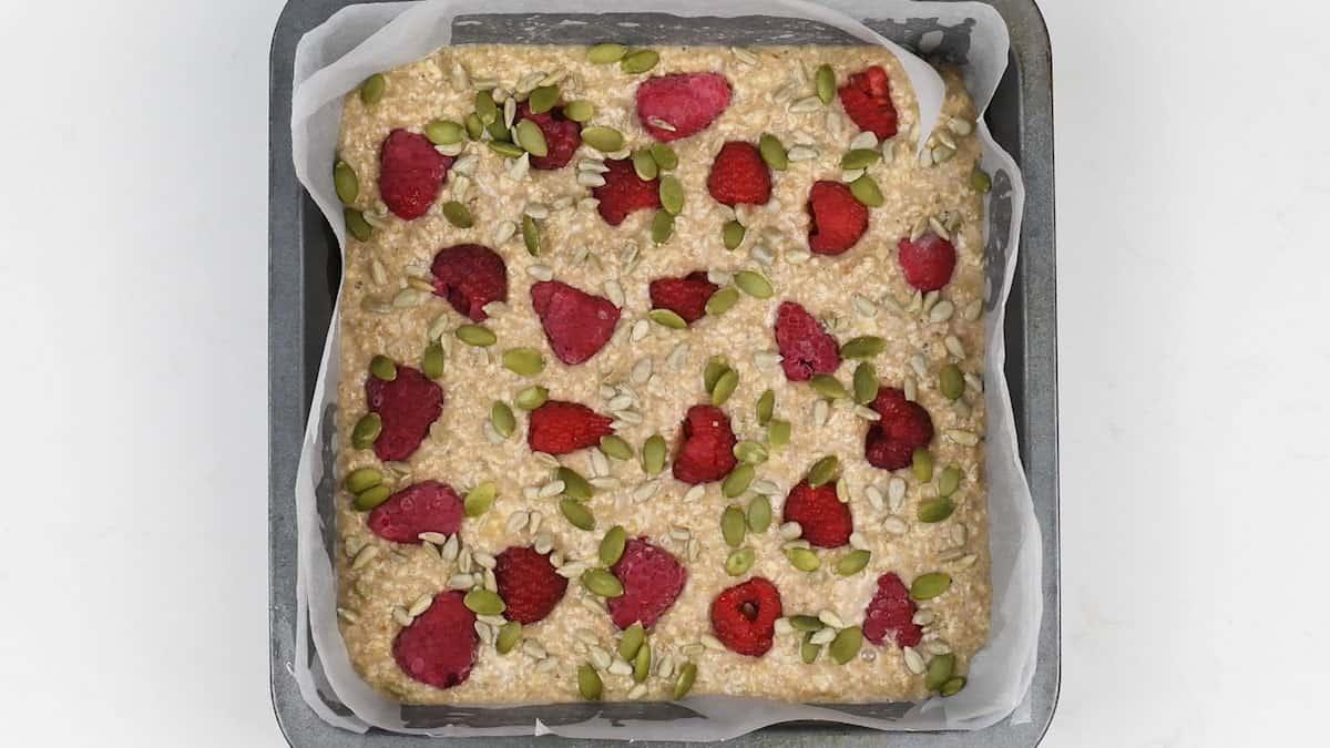 A square baking pan lined with parchment paper, filled with Healthy Baked Oats mixture, topped with fresh raspberries, sunflower seeds, and pumpkin seeds.