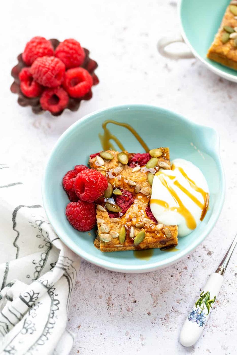 A blue bowl with two Healthy Baked Oats bars topped with seeds, a spoonful of yogurt drizzled with honey, and fresh raspberries; extra raspberries and a cloth napkin are nearby.