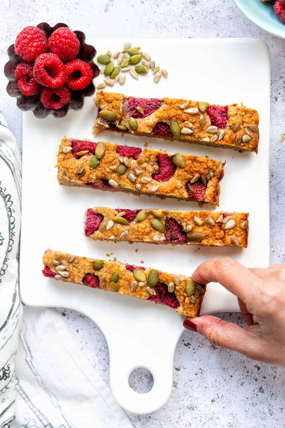 Four Healthy Baked Oats raspberry bars topped with seeds are arranged on a white cutting board, with a small dish of fresh raspberries nearby. A hand is reaching in to pick up one of the bars.