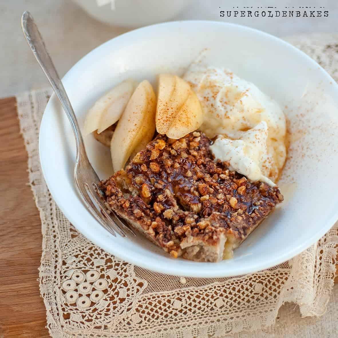 A white bowl with a piece of Healthy Baked Oats, sliced apples, and a dollop of yogurt or cream, sprinkled with cinnamon, on a lace napkin with a fork.