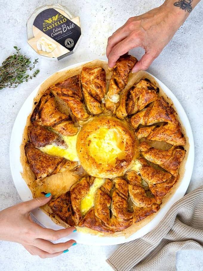 Two hands reach for pieces of a golden, flaky Brie in Puff Pastry Tarte Soleil with melty brie cheese in the center, served on a white plate. A wheel of Castello brie and fresh herbs rest nearby on a light surface.