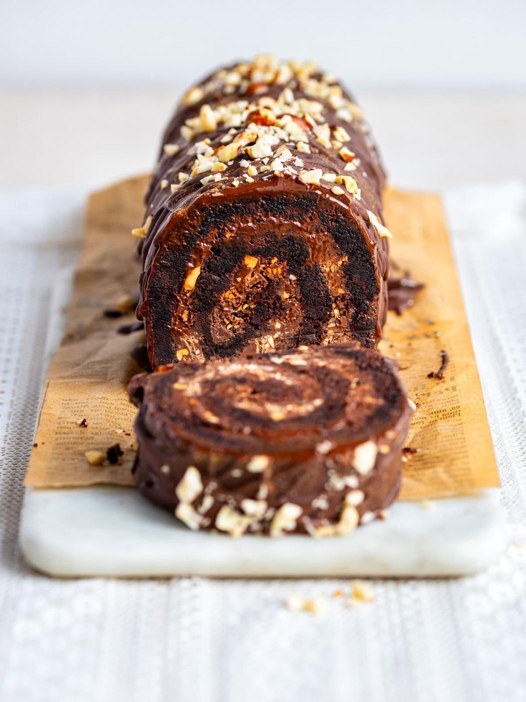 A chocolate Brownie Yule log with a creamy filling, topped with chopped nuts and drizzled chocolate, sits sliced on a wooden board with a marble base.