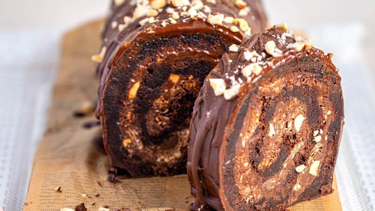 A chocolate brownie roll is topped with chopped nuts and sliced on a parchment-lined marble board. The cake features a rich swirl pattern inside and is elegantly drizzled with chocolate.