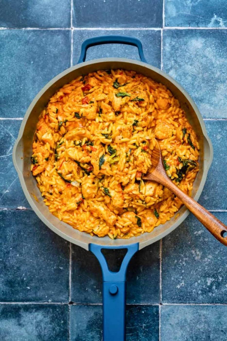 A pan filled with creamy, orange-colored Cajun chicken orzo—tender chicken chunks, rice, and leafy greens—stirred with a wooden spoon, sits on a dark blue tiled surface.