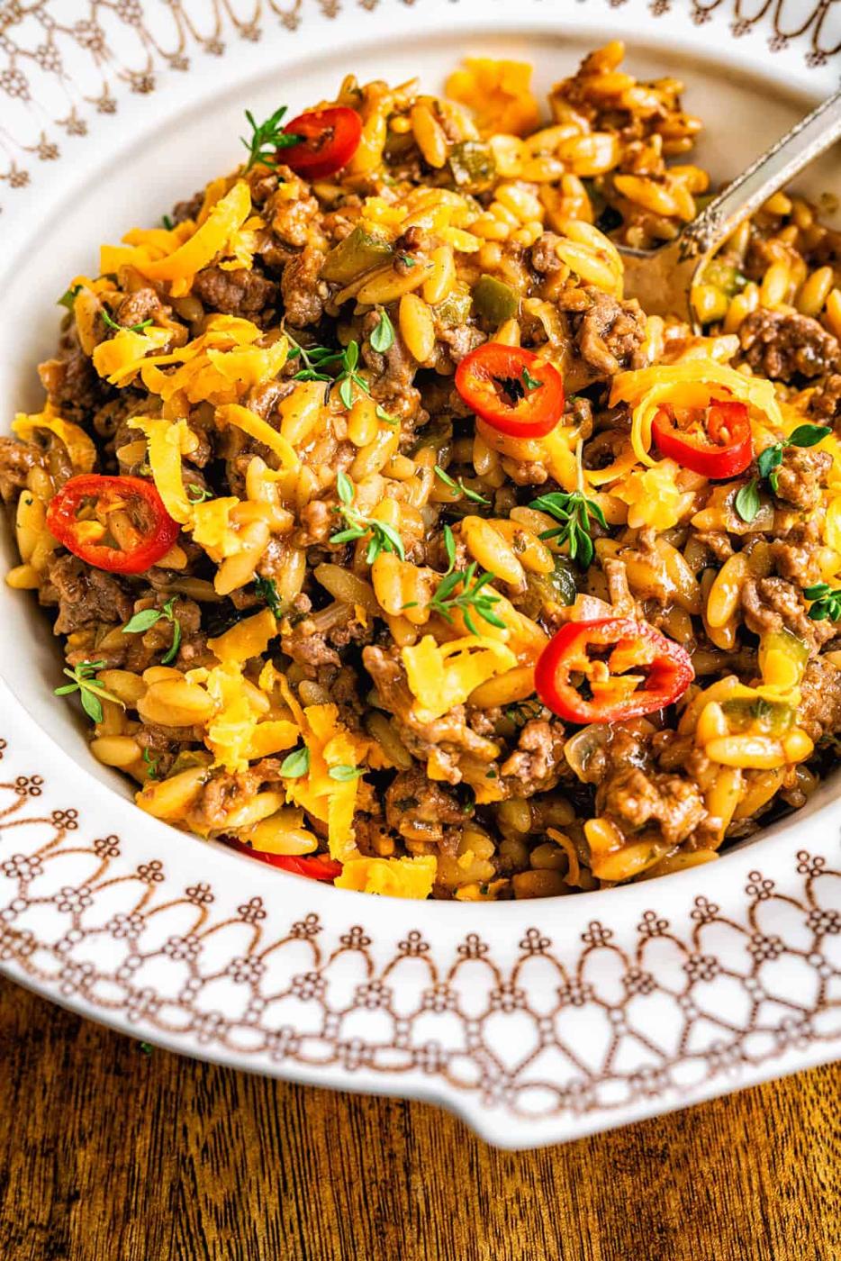A bowl of cheesy Cajun dirty orzo with ground beef, topped with sliced red chili peppers and fresh herbs, served in a decorative dish on a wooden table.
