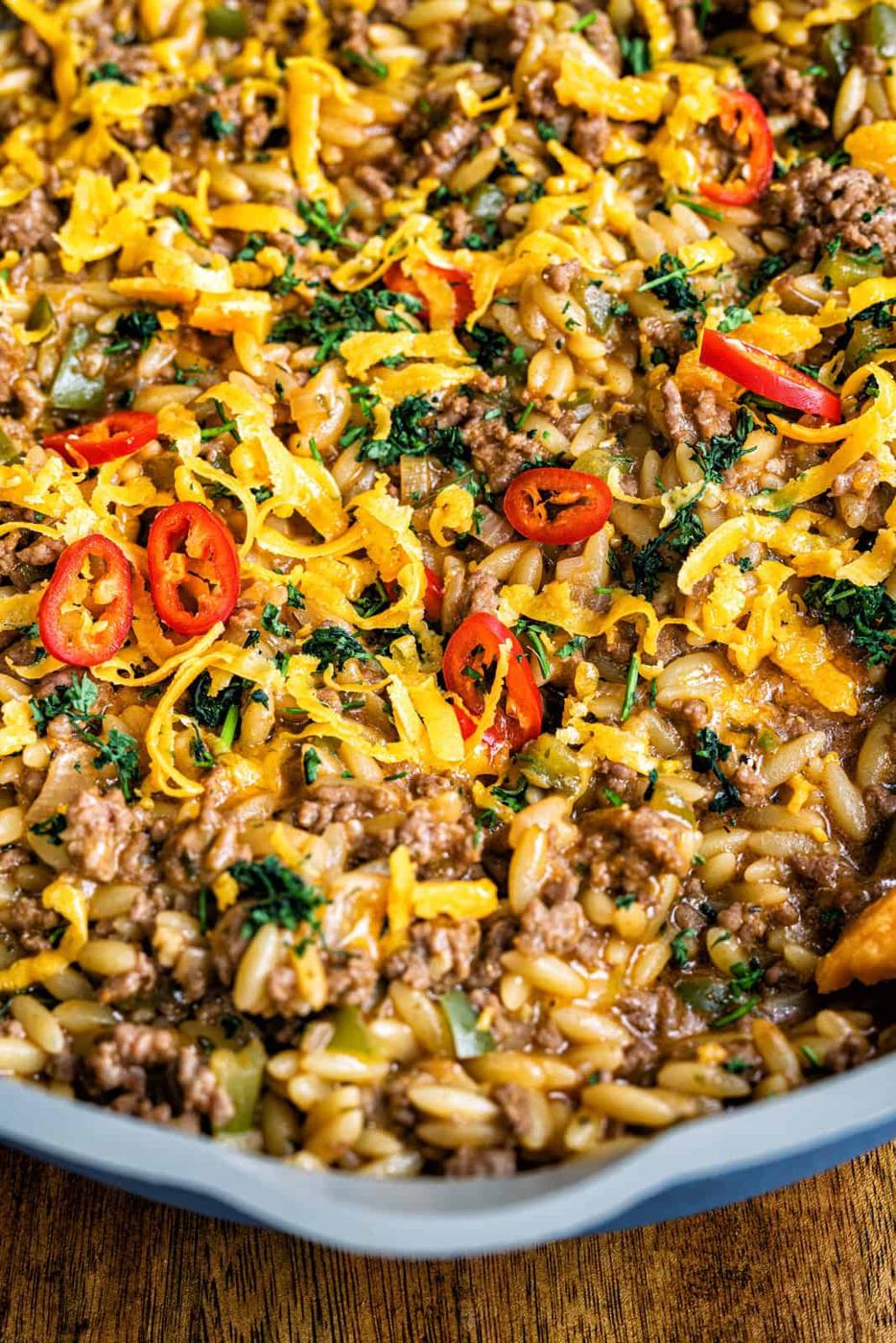 A close-up of a skillet filled with cheesy cajun dirty orzo and ground beef, garnished with chopped parsley and sliced red chili peppers.