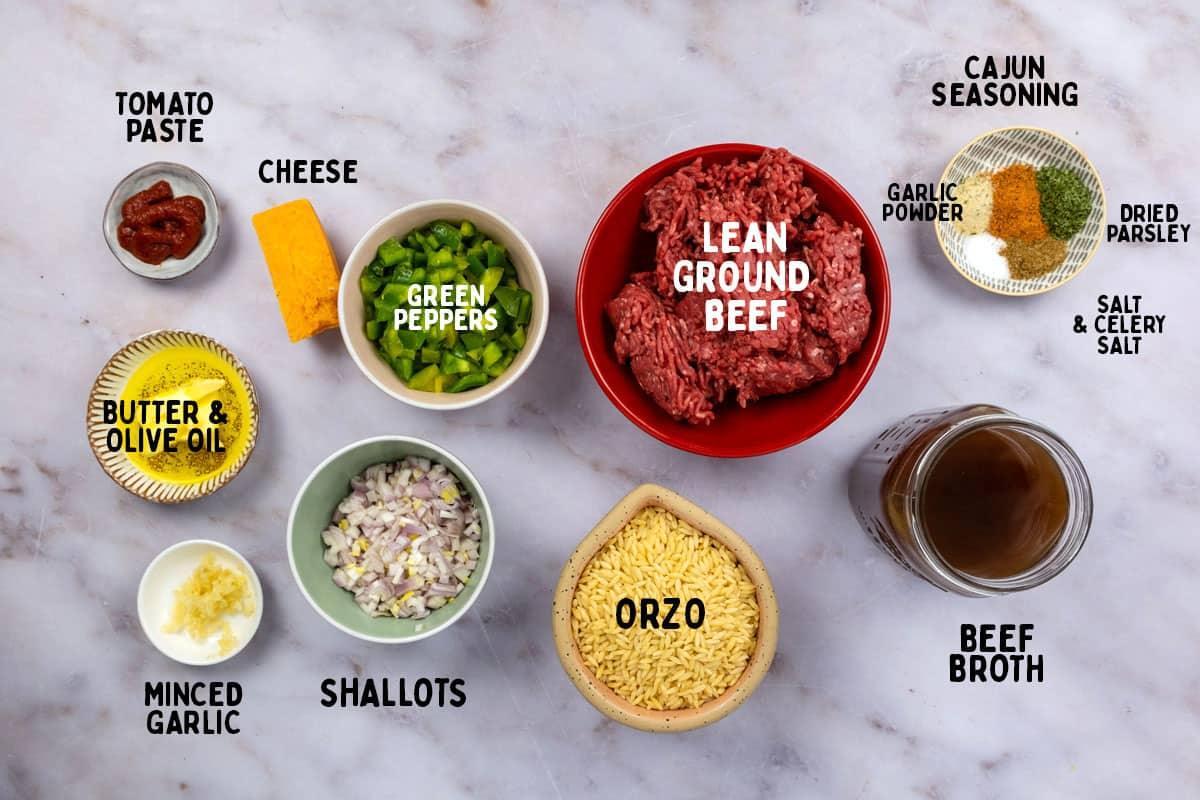 Ingredients for Cajun dirty orzo laid out on a marble surface, including labeled bowls of lean ground beef, orzo, beef broth, shallots, minced garlic, butter & olive oil, tomato paste, cheese, green peppers, and assorted seasonings.