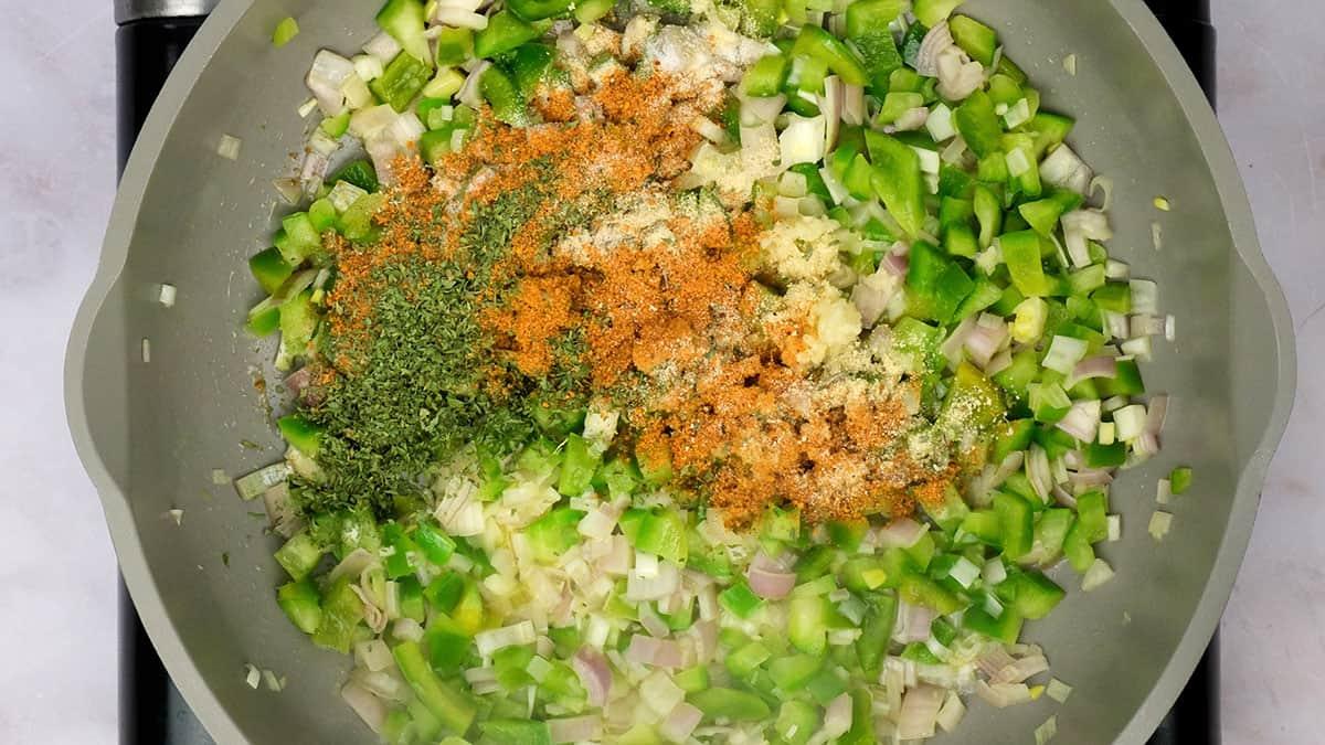 Chopped green bell peppers, onions, and leeks are being sautéed in a pan with cajun dirty orzo, various spices, and dried herbs sprinkled on top.