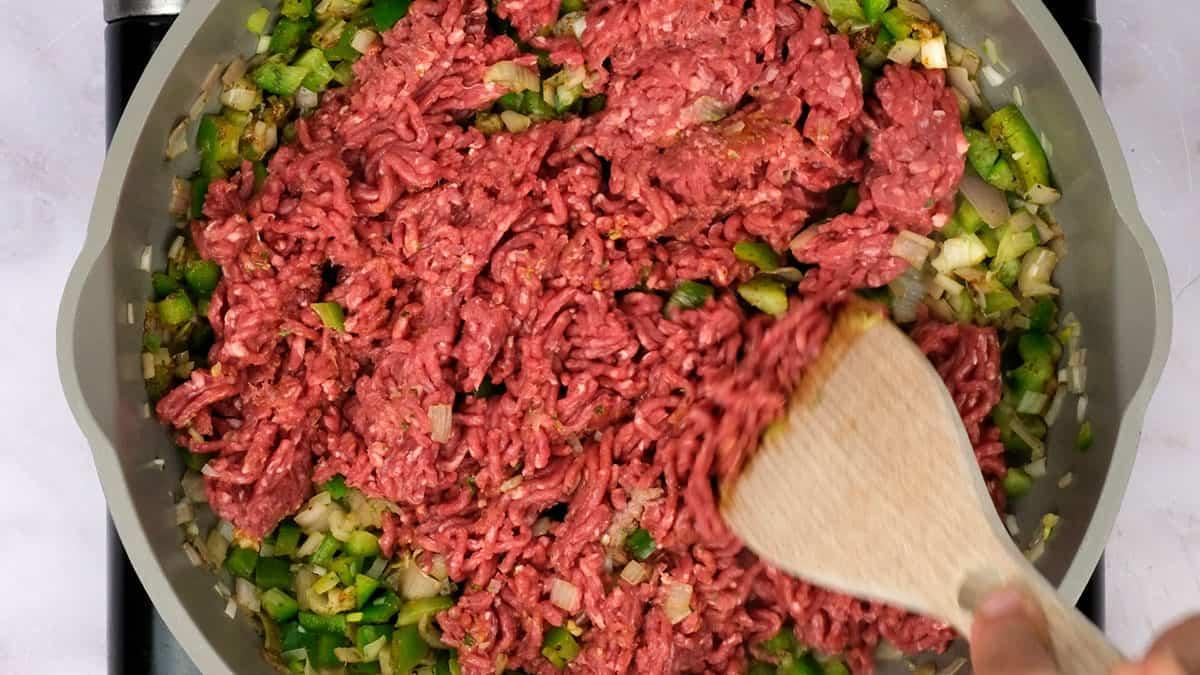 A hand stirs raw ground beef into a pan with chopped green bell peppers and onions using a wooden spatula, starting the base for a flavorful cajun dirty orzo dish.
