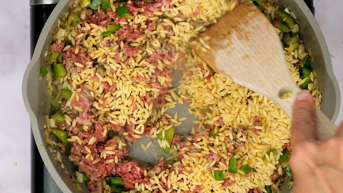 A hand stirs cajun dirty orzo, ground meat, and chopped green bell peppers in a frying pan using a wooden spatula.
