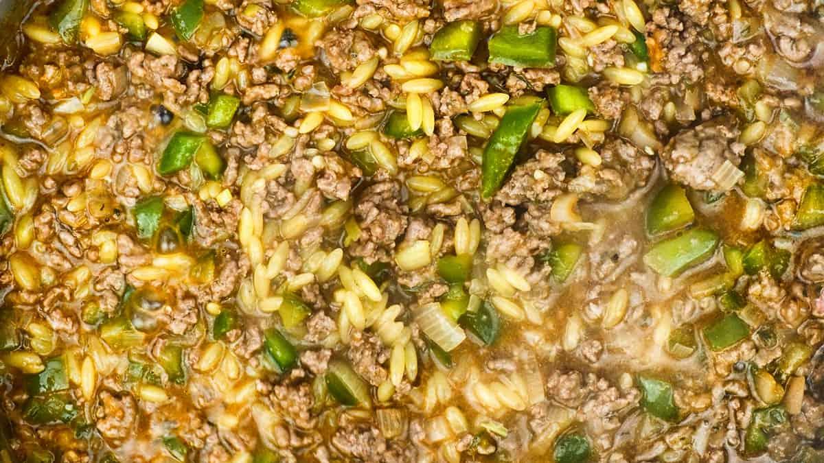A close-up of a skillet filled with ground beef, chopped green bell peppers, rice, onions, and a savory sauce, all mixed together and simmering in the style of Cajun dirty orzo.