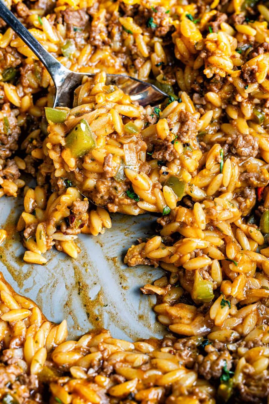 Close-up of a cheesy Cajun dirty orzo skillet with ground beef, diced green bell peppers, fresh herbs, and a spoon scooping up a savory portion from the pan.