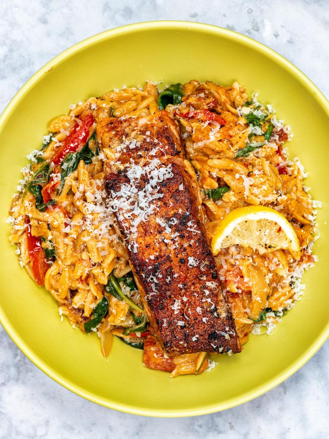 A yellow plate with creamy orzo, spinach, and red bell peppers, topped with a blackened salmon fillet, grated cheese, and a slice of lemon.