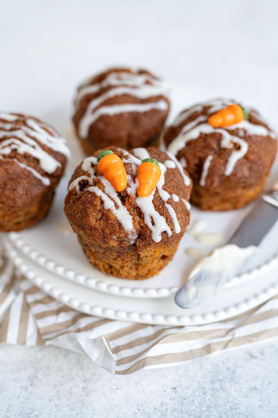 Four carrot cake muffins with white icing drizzled on top, two decorated with small fondant carrots, are arranged on a white plate next to a butter knife on a striped cloth.