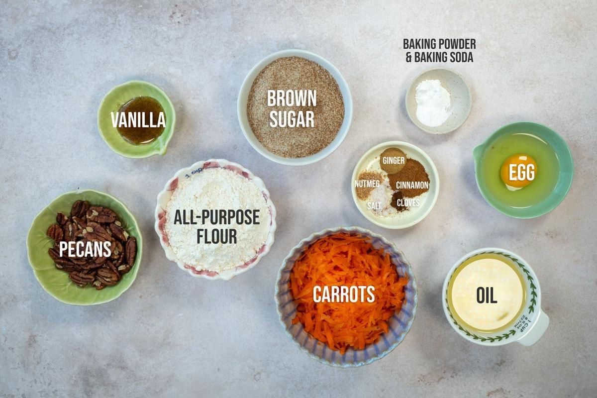 Top-down view of bowls containing labeled carrot cake ingredients: vanilla, brown sugar, egg, oil, shredded carrots, all-purpose flour, pecans, and a bowl with baking powder, baking soda, and spices.