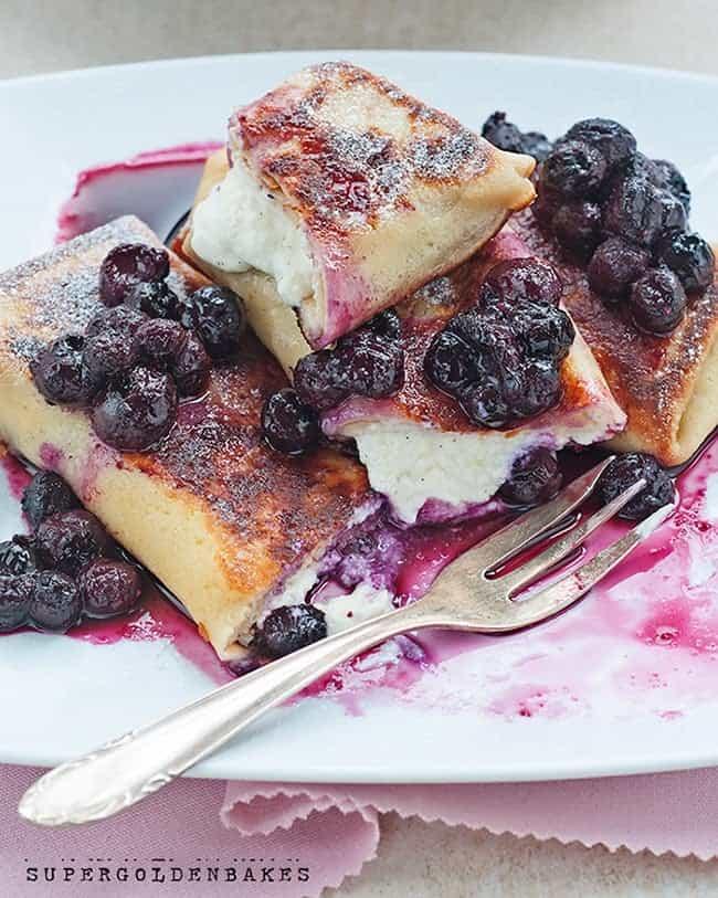 Three golden-brown cheese blintzes are topped with sweet, juicy blueberries and served on a white plate with a fork. Purple blueberry sauce is drizzled around the cheese blintz, creating a vibrant and appetizing presentation.