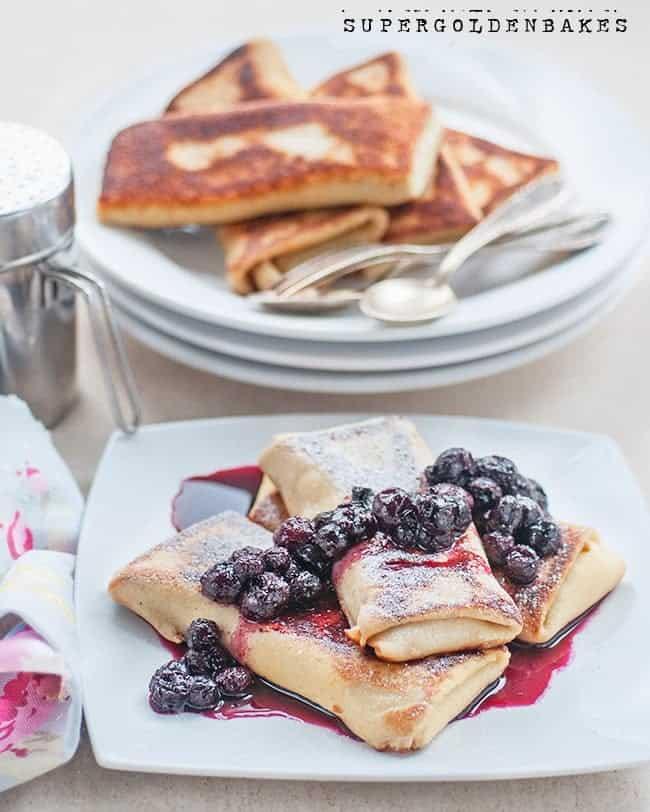 Three blitz filled and folded into rectangles on a white plate, topped with blueberries and berry sauce, with powdered sugar sprinkled on top. In the background, more crepes and utensils are stacked on plates.