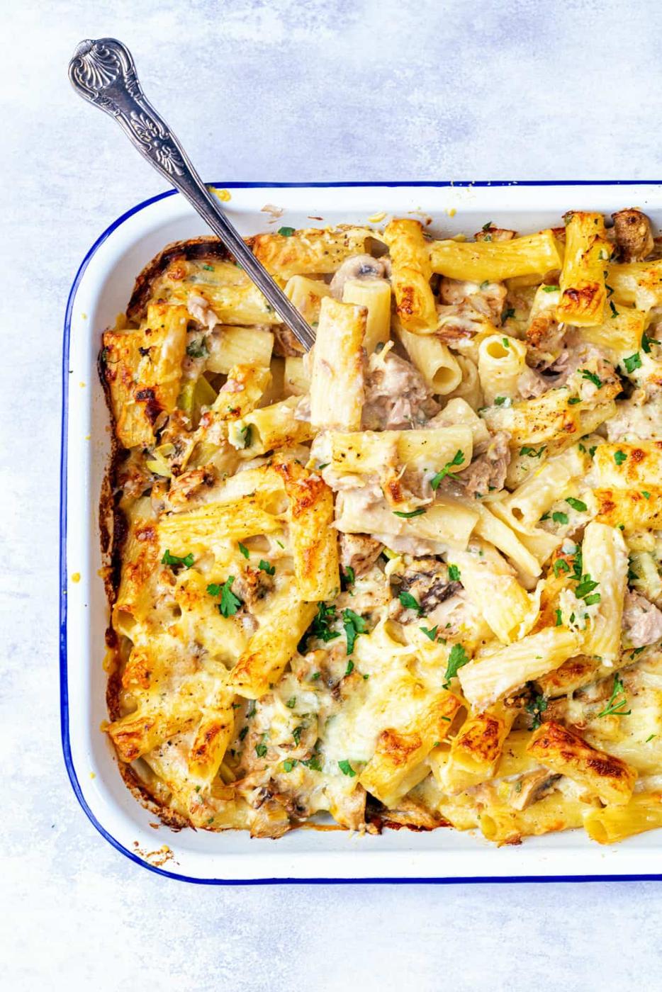 A Cheesy Tuna Pasta Bake casserole with rigatoni, mushrooms, and cheese, garnished with fresh herbs in a white dish, with a serving spoon scooping out a portion.