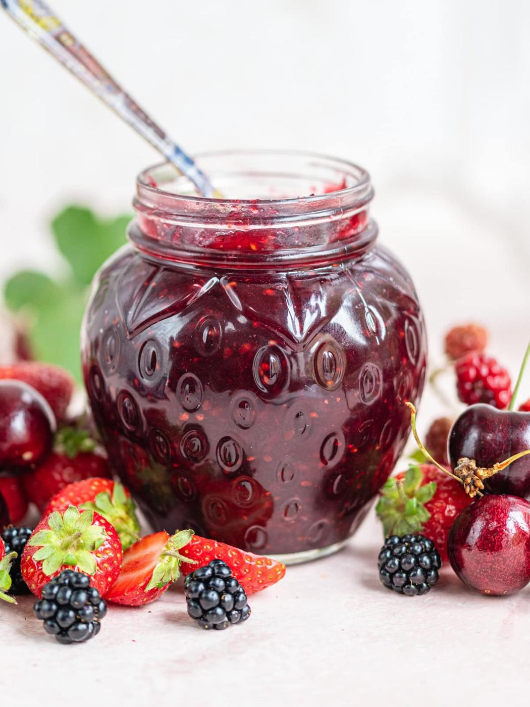 A glass jar filled with berry jam sits on a table surrounded by fresh strawberries, cherries, and blackberries, with a spoon inside the jar.
