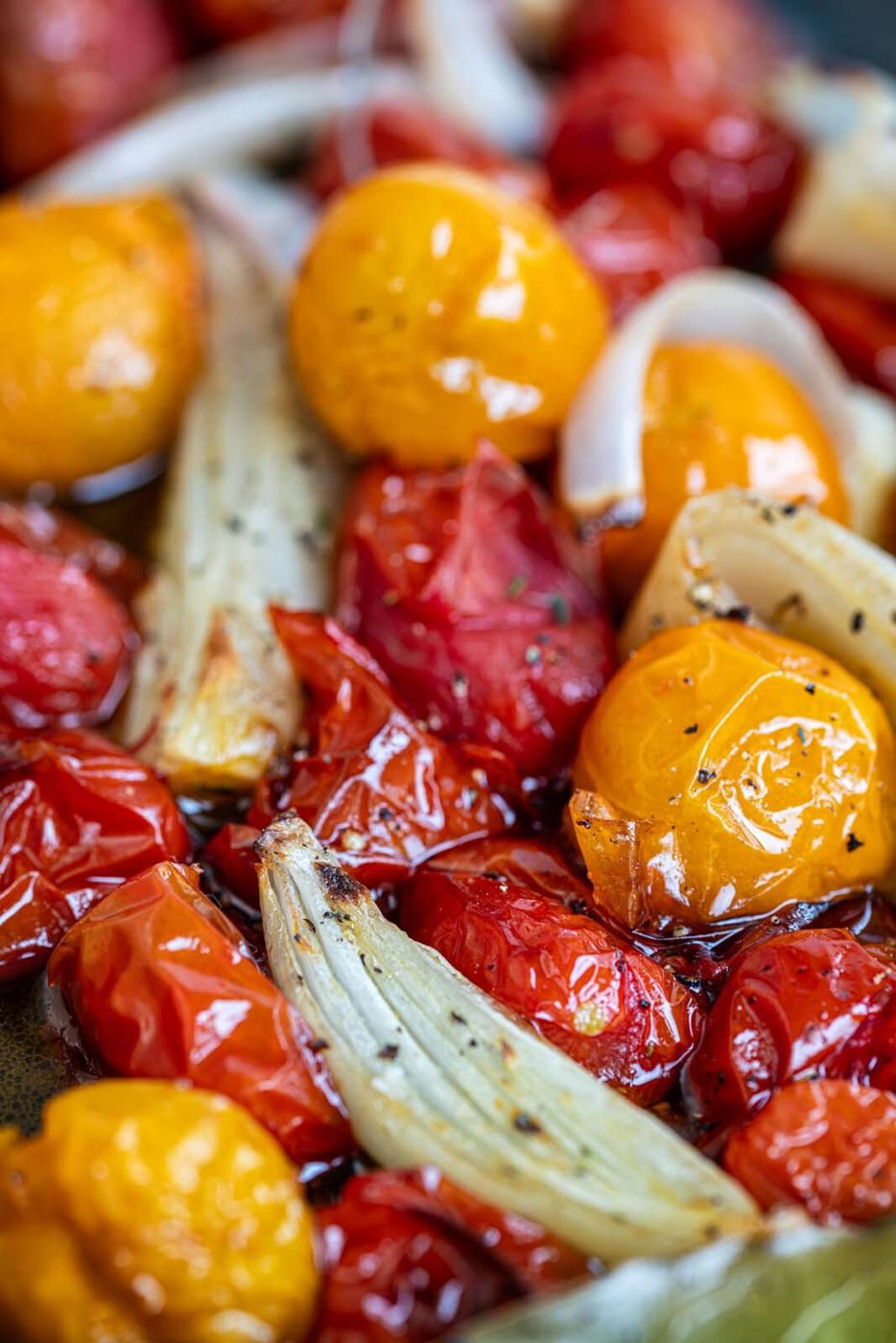 Close-up of roasted cherry tomatoes in red and yellow, mixed with roasted onion slices, glistening with olive oil and sprinkled with cracked black pepper.