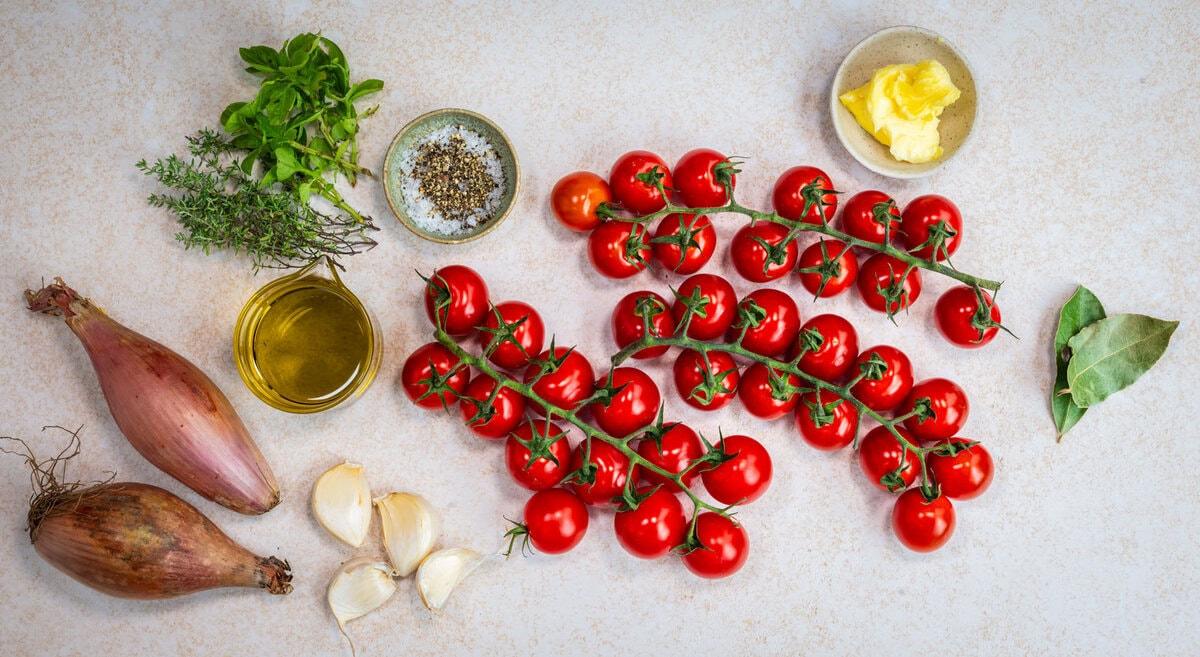A top-down view of cooking ingredients, including vine tomatoes, shallots, garlic cloves, olive oil, fresh herbs, pepper, bay leaves, and a small dish of butter, arranged neatly on a light surface.