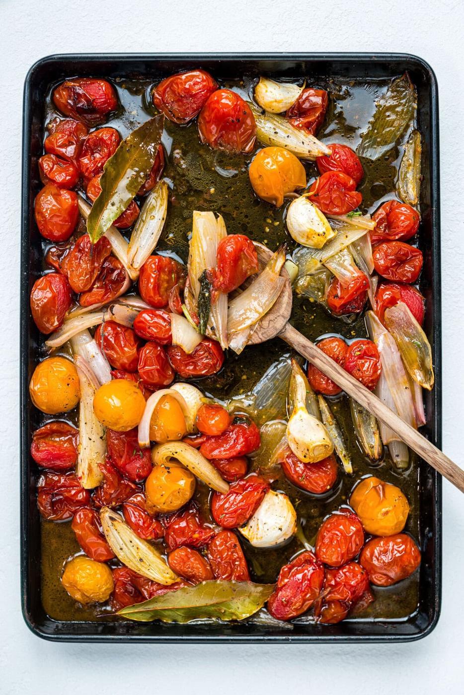 A baking tray filled with roasted cherry tomatoes, shallots, garlic cloves, and bay leaves, all coated in olive oil, with a wooden spoon resting on the tray.