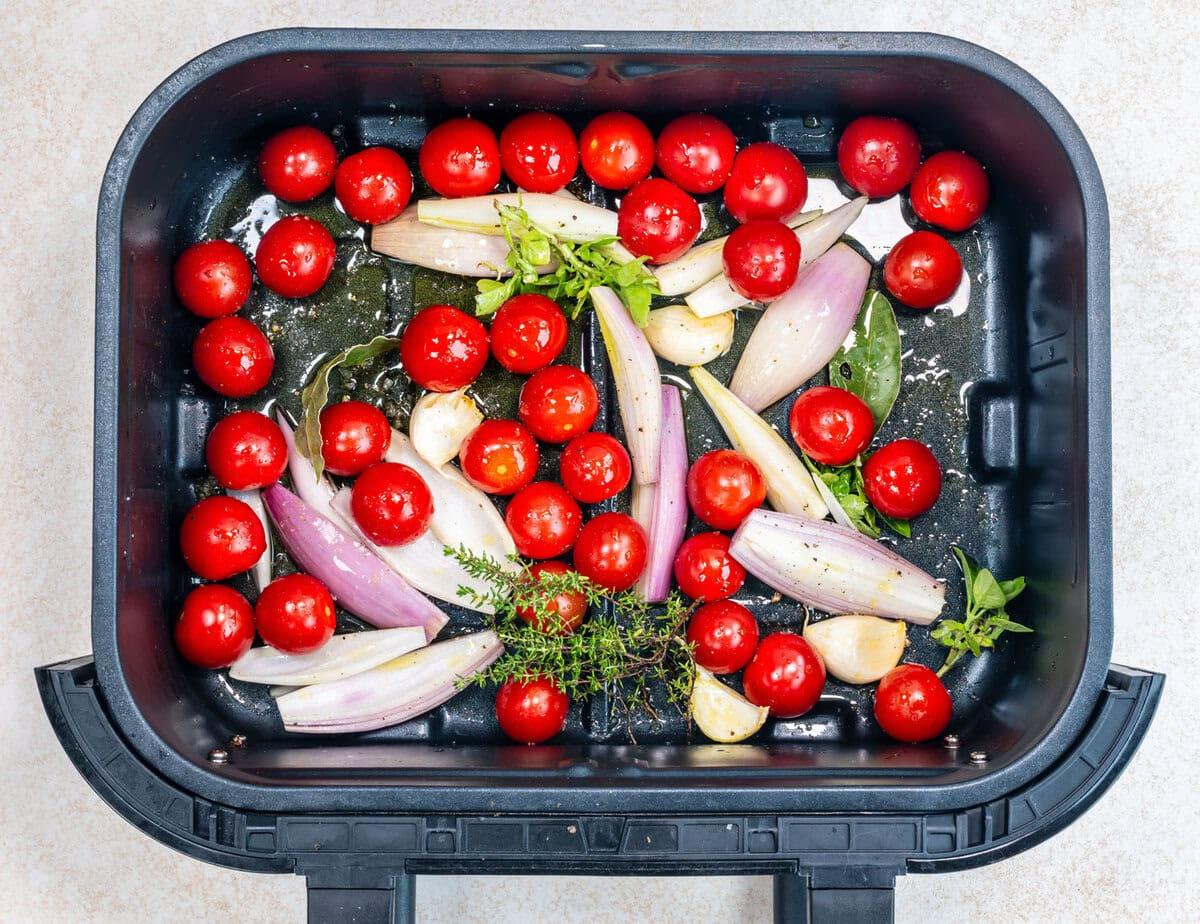 Cherry tomatoes, shallots, garlic cloves, fresh herbs, and seasonings are arranged in a black air fryer basket, ready for roasting.