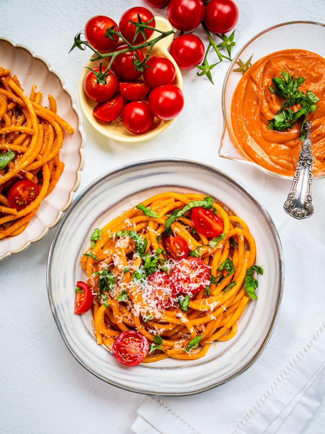 A bowl of pasta topped with fresh cherry tomato sauce, cherry tomatoes, grated cheese, and fresh herbs sits on a white table beside a plate of pasta, a bowl of whole cherry tomatoes, and a bowl of tomato sauce with a spoon.