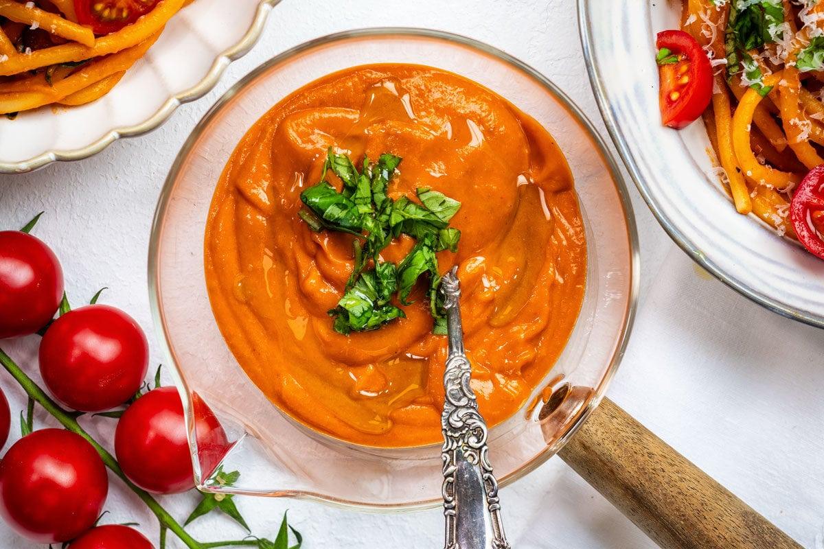 A glass bowl of creamy tomato sauce topped with chopped basil is surrounded by fresh cherry tomatoes and plates of pasta garnished with grated cheese and basil on a white surface.