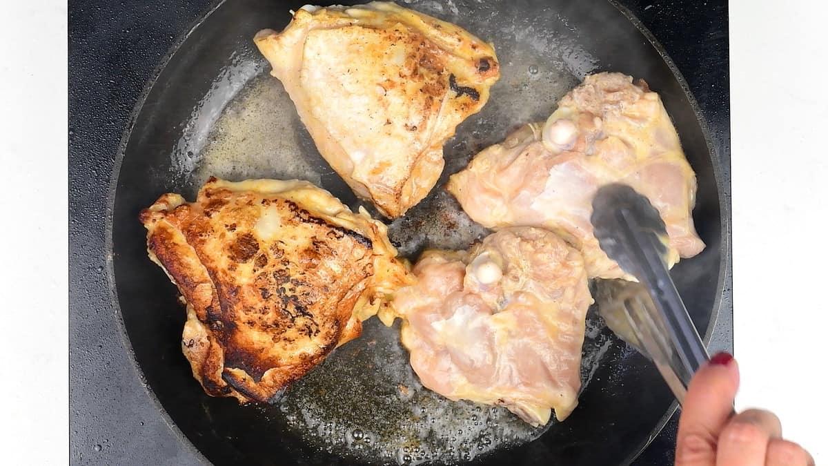 A hand uses tongs to cook four bone-in chicken thighs in a skillet for chicken adobo; two pieces are browned, while two remain raw. Steam rises from the hot pan.