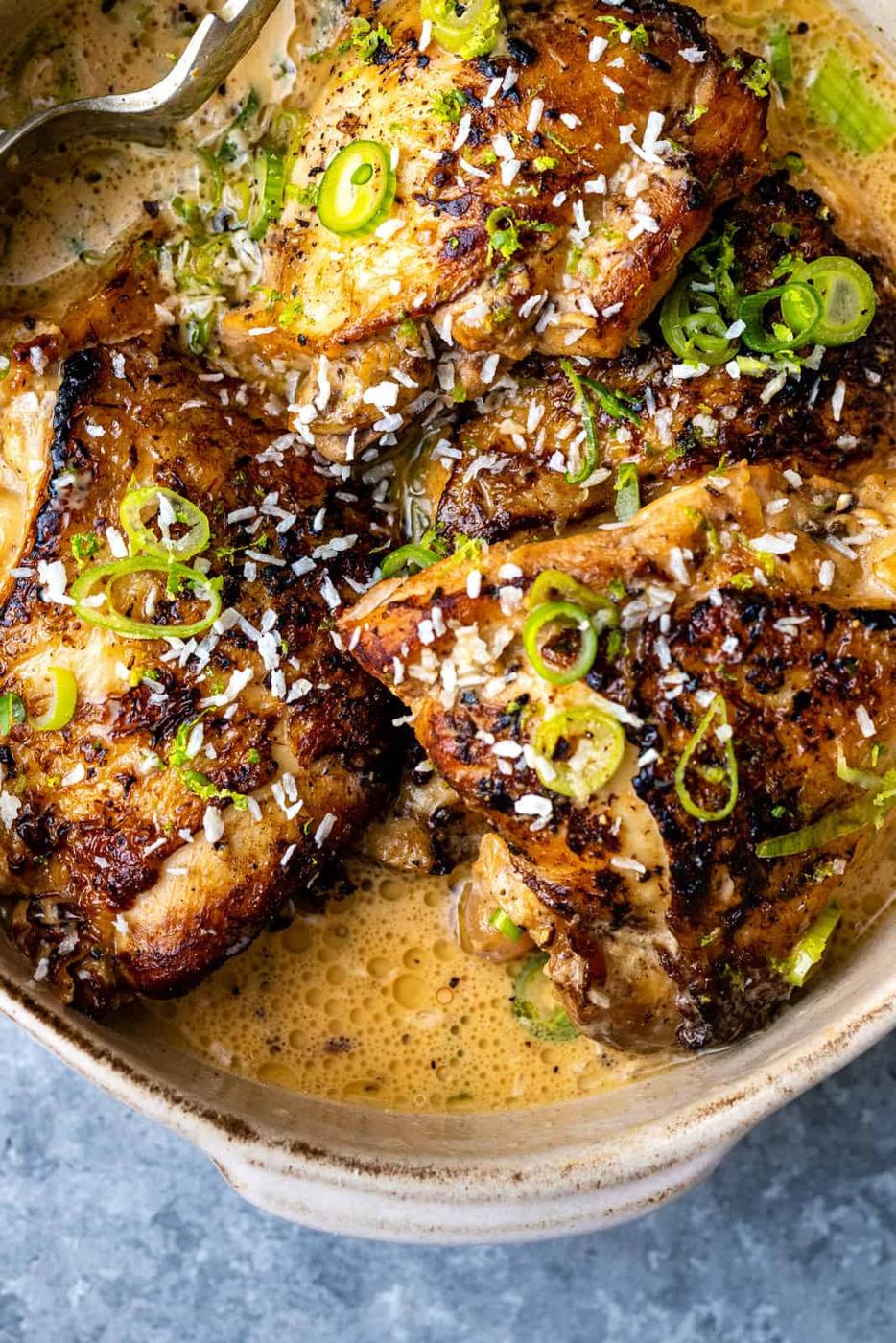 Golden-brown chicken thighs, inspired by chicken adobo, simmer in a creamy sauce and are garnished with shredded coconut and sliced green onions in a ceramic dish on a gray tabletop.