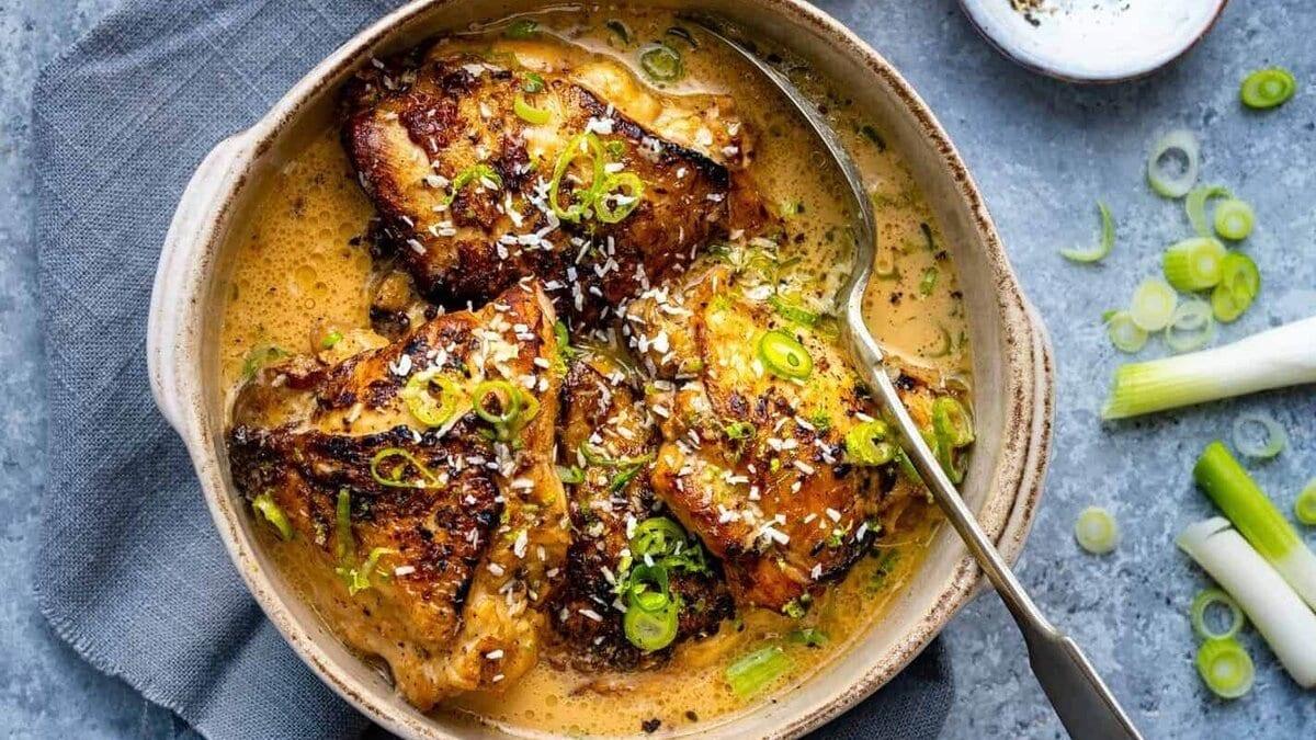A bowl of golden-brown slow cooker chicken thighs in a creamy sauce, garnished with sliced green onions and herbs. A spoon rests in the bowl, which sits on a gray napkin on a stone surface.