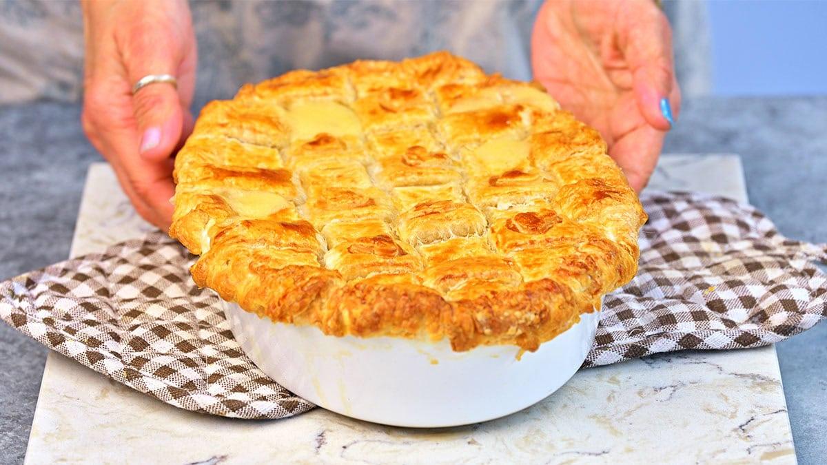 A person places a freshly baked pie with a golden, flaky lattice crust on a checkered cloth, the pie resting in a white ceramic dish on a marbled surface.