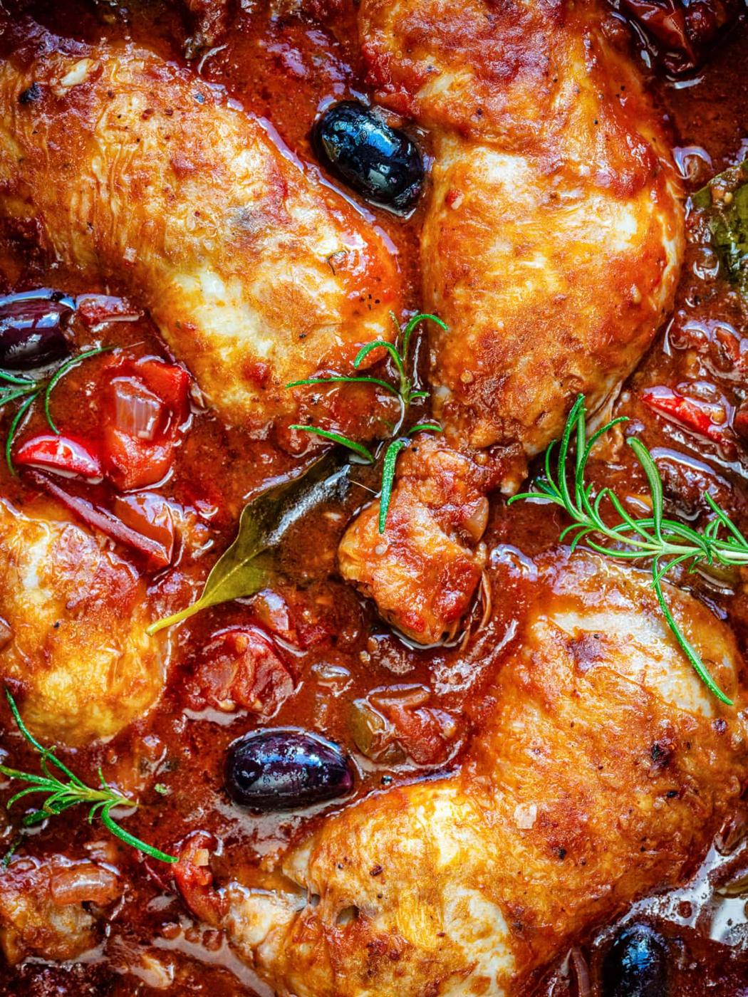 Golden-brown chicken pieces in a rich tomato Cacciatore sauce with black olives, cherry tomatoes, bay leaves, and sprigs of fresh rosemary. The dish appears savory and freshly cooked.