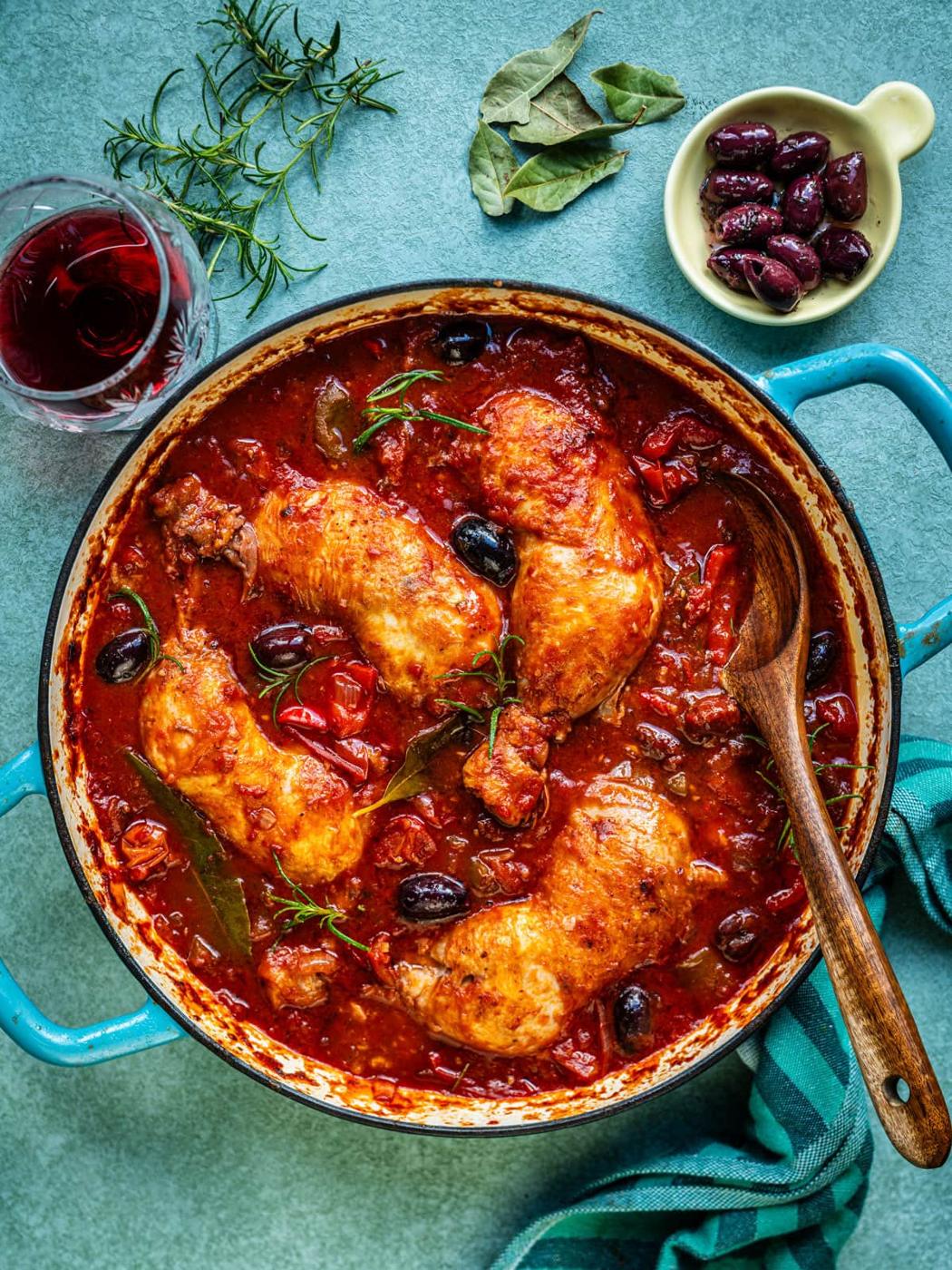 A pot of braised chicken cacciatore in a rich tomato sauce with black olives and herbs, wooden spoon inside, surrounded by a glass of red wine, fresh herbs, and a small bowl of olives on a blue surface.