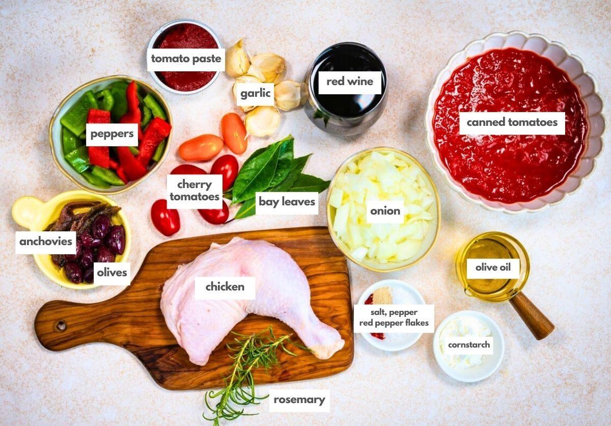 Ingredients for a Chicken Cacciatore recipe are arranged on a table, each labeled: chicken, rosemary, olives, anchovies, peppers, cherry tomatoes, tomato paste, garlic, bay leaves, onion, canned tomatoes, red wine, olive oil and seasonings.