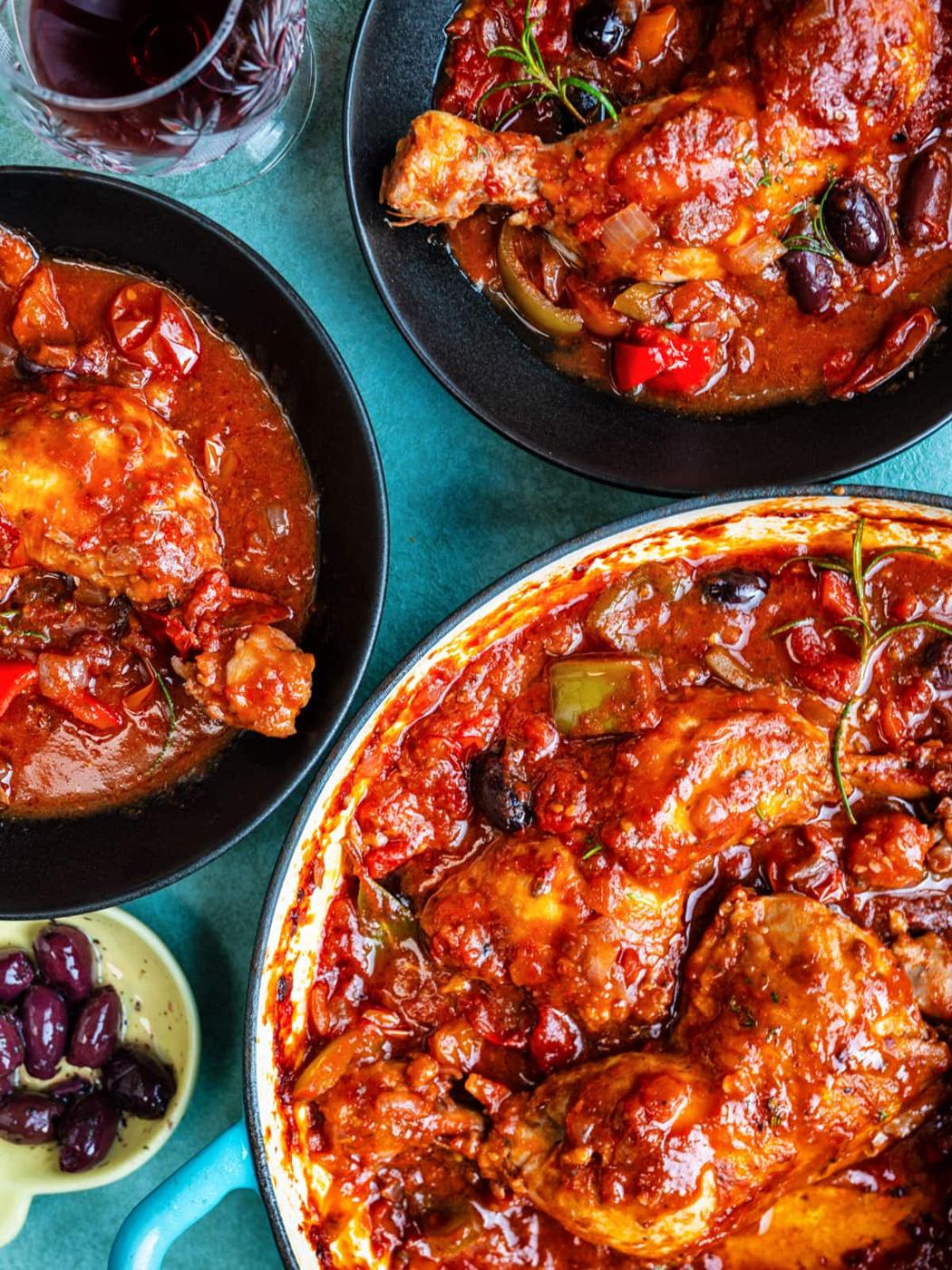 A large skillet and two plates filled with chicken Cacciatore in a rich tomato sauce with olives, peppers, and herbs. A small bowl of olives and a glass of red wine sit nearby on a blue tabletop.