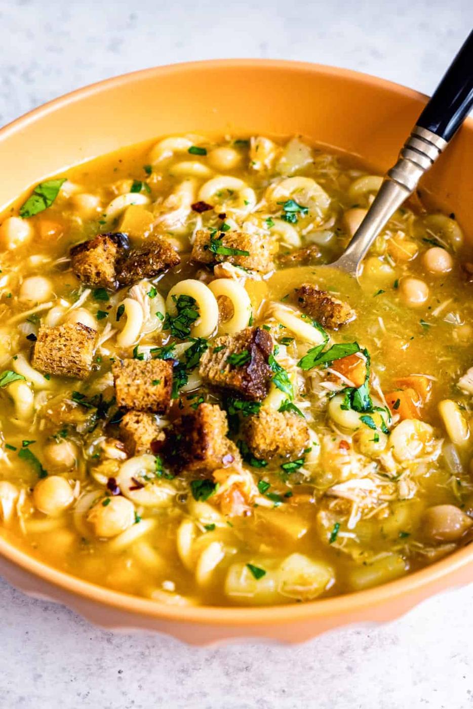 A bowl of Chicken Carcass Soup with pasta, chickpeas, chopped herbs, and croutons, served in an orange bowl with a spoon.