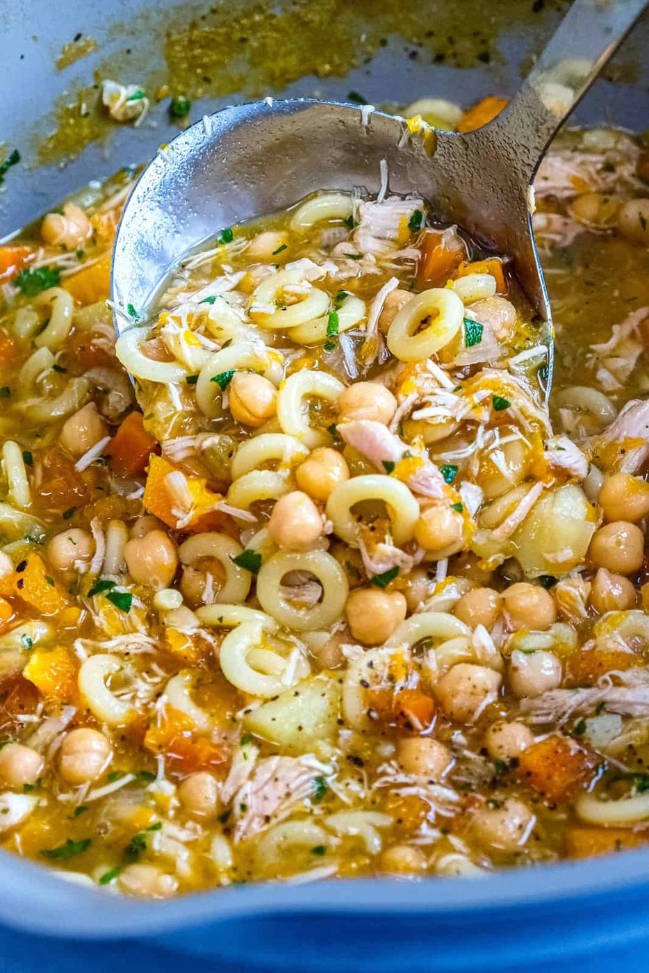 A ladle scoops hearty Chicken Carcass Soup with circular pasta, chickpeas, shredded chicken, diced carrots, potatoes, and herbs in a rich broth, all simmering in a large pot.