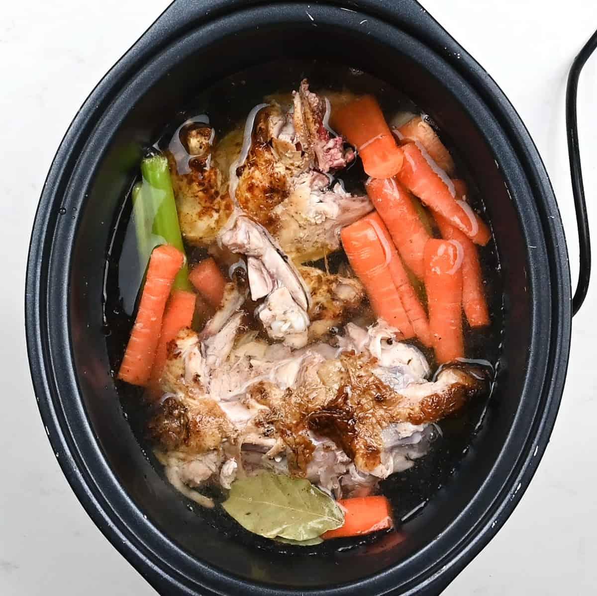 A slow cooker filled with chicken bones, chopped carrots, celery, and a bay leaf submerged in water, preparing to make delicious Chicken Carcass Soup.