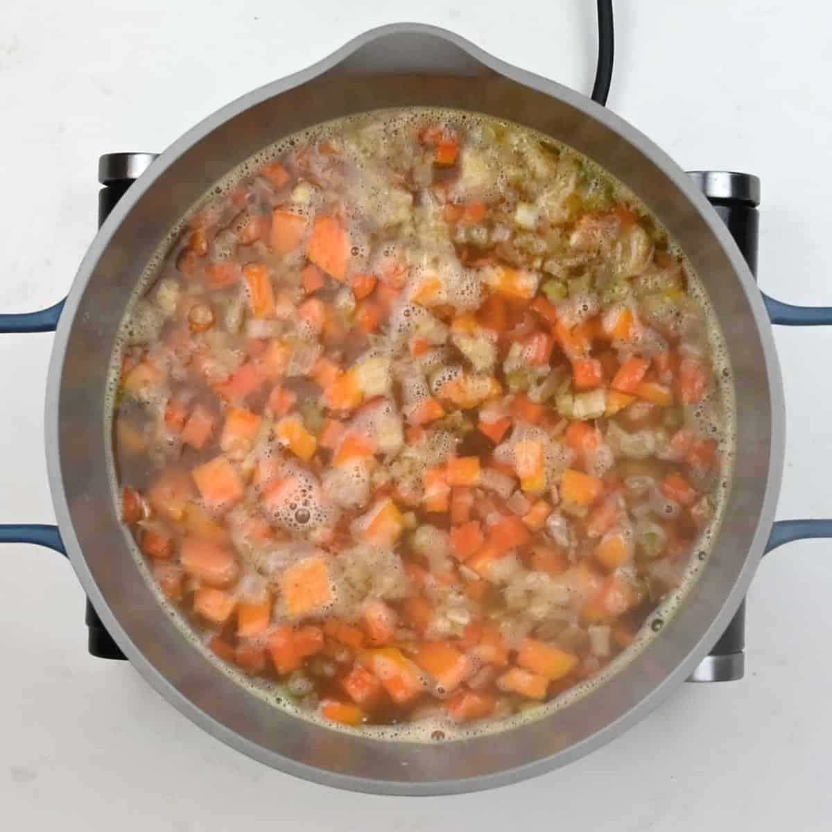 A top-down view of a pot on a stove filled with chopped carrots, onions, and celery simmering in bubbling broth for a hearty Chicken Carcass Soup.