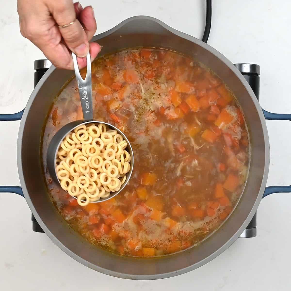 A hand holds a measuring cup filled with ring-shaped pasta above a pot of simmering Chicken Carcass Soup with diced carrots and a rich brown broth.