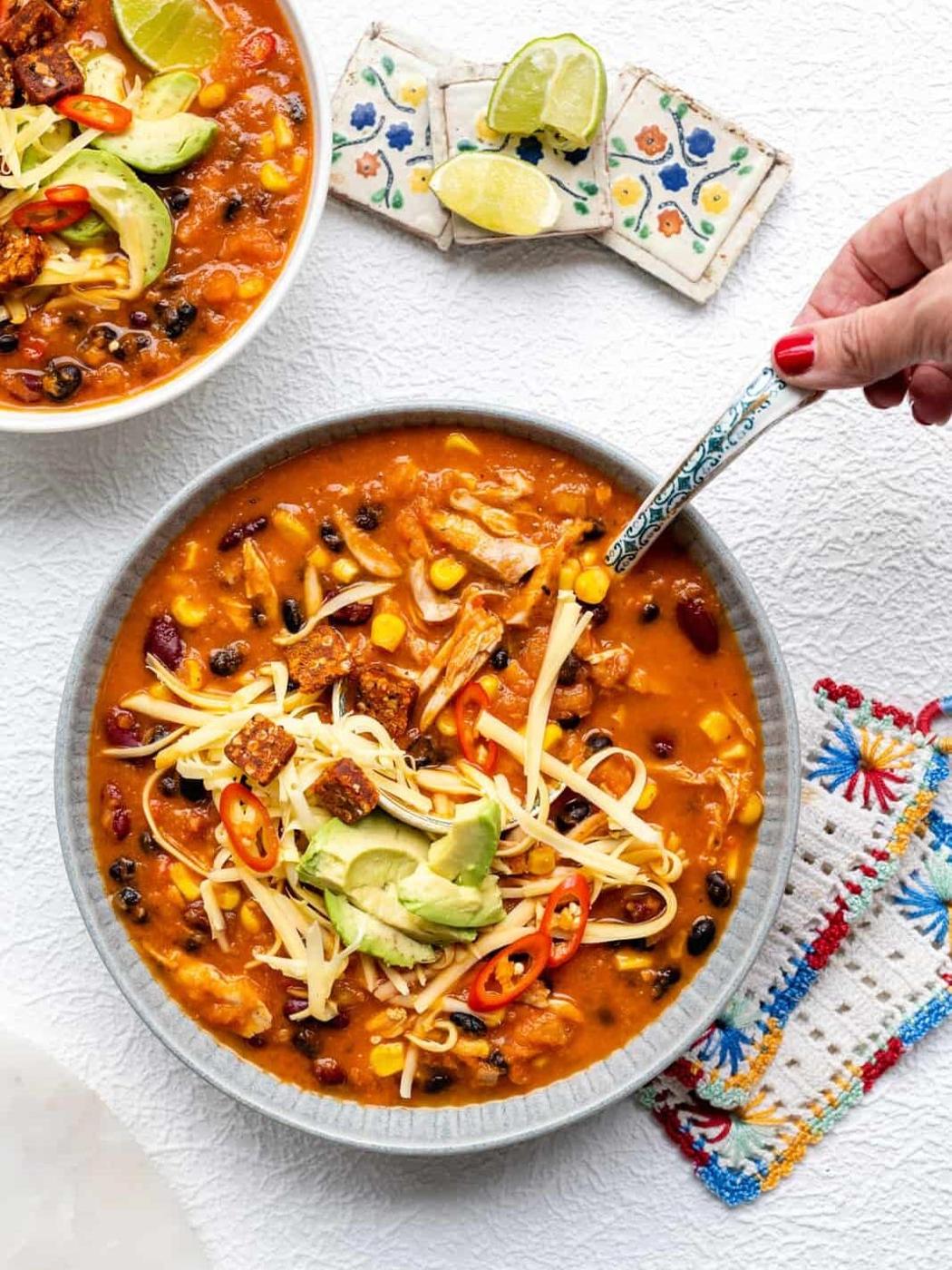 A bowl of enchilada chicken soup topped with shredded cheese, avocado slices, tortilla strips, and chili peppers; a hand holds a spoon. Another bowl of soup and lime wedges are visible in the background.