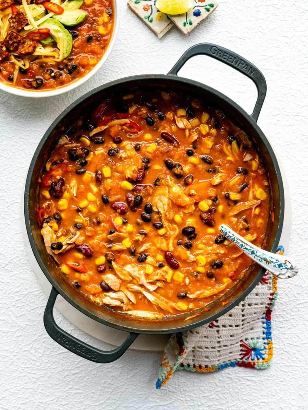 A pot of hearty chicken chili with beans, corn, tomatoes, and shredded chicken in a rich, orange-red sauce, shown with a spoon and a bowl of chili topped with avocado and tortilla strips nearby.