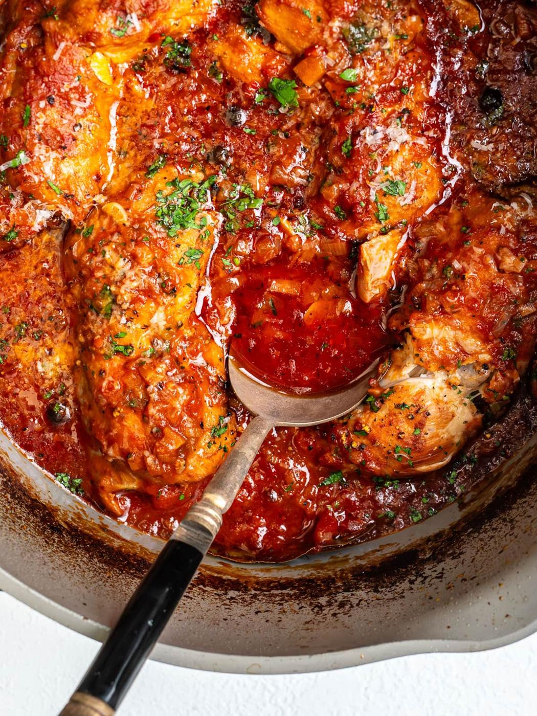 A close-up of chicken leg quarters simmering in a rich tomato sauce, garnished with chopped herbs, with a spoon lifting some of the sauce in a large pan.