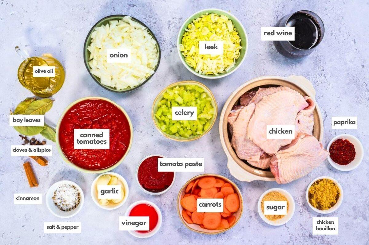 Top-down view of labeled ingredients for Greek Chicken Kokkinisto on a table, featuring raw chicken in tomato sauce, canned tomatoes, onion, leek, celery, garlic, carrots, tomato paste, red wine, olive oil, spices, vinegar, and chicken bouillon.