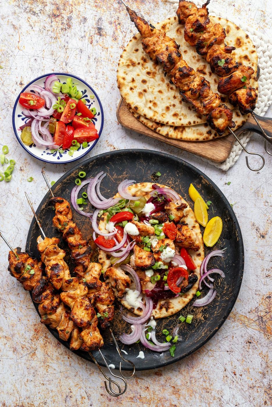 Chicken Kebabs on flatbreads, garnished with sliced red onions, tomatoes, green onions, lemon wedges, and served with a side salad in a small bowl. The food is beautifully arranged on rustic plates and boards.