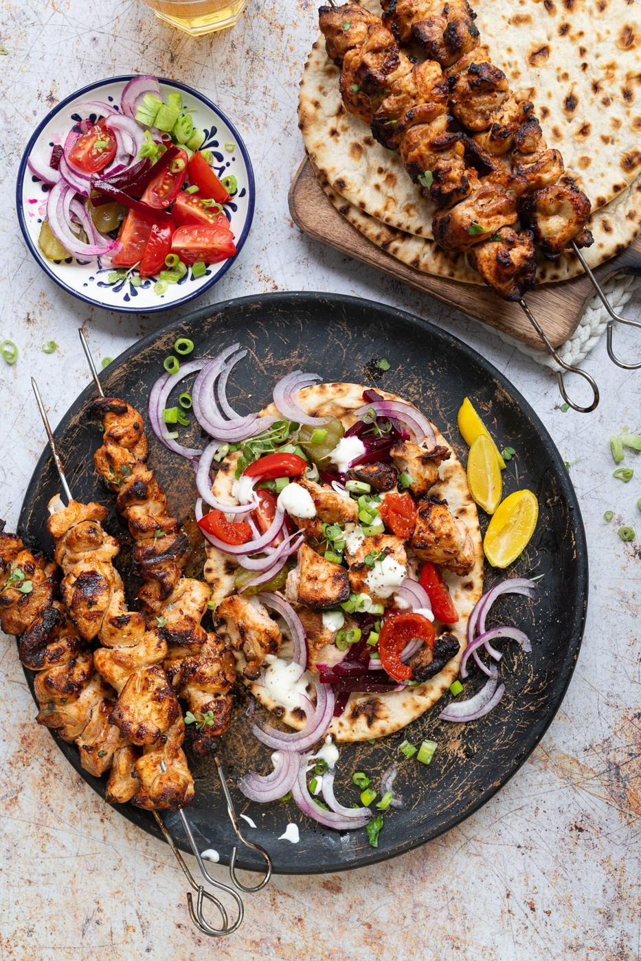 Chicken Kebabs skewers on a platter with flatbread, sliced onions, tomatoes, green onions, lemon wedges, and a side salad of tomatoes and onions. The food is attractively arranged on a rustic background.