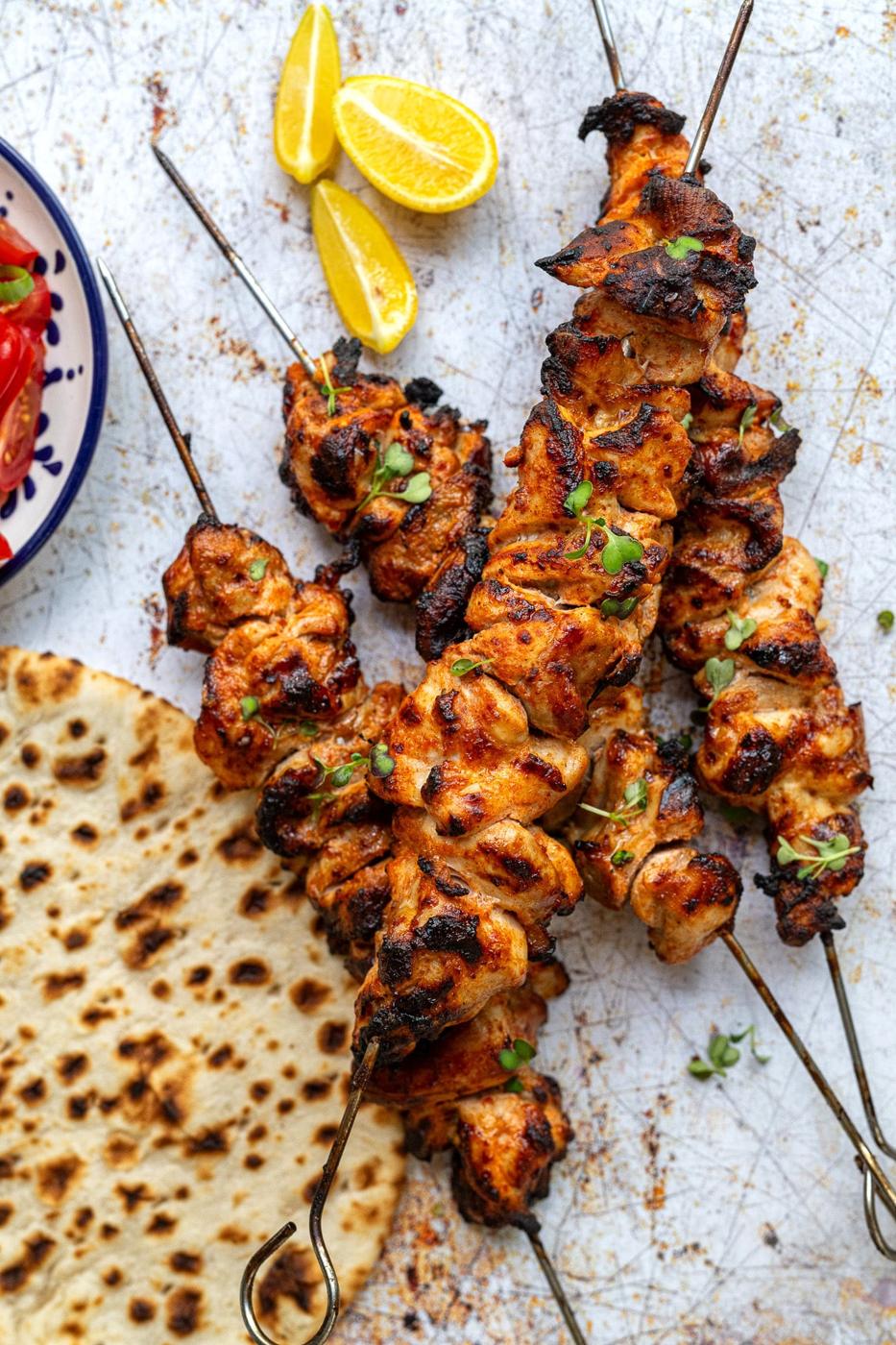 Grilled chicken kebabs with char marks, garnished with herbs, served alongside flatbread and lemon wedges on a rustic white surface.